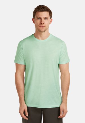 Man die een lichtgroen T-shirt met korte mouwen en ronde hals draagt, en donkere broek, staand tegen een effen witte achtergrond.