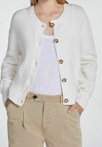 Vit texturerad cardigan med rund halsringning, prydd med guldknappar fram. Bärs över en enkel vit topp och beige byxor.