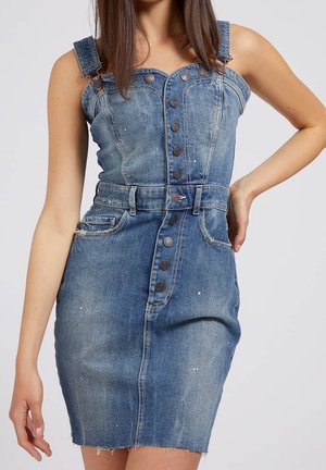 Kvinde iført ærmeløs blå denimkjole med knapper foran, bæltestropper og forlommer, står med den ene hånd på hoften.