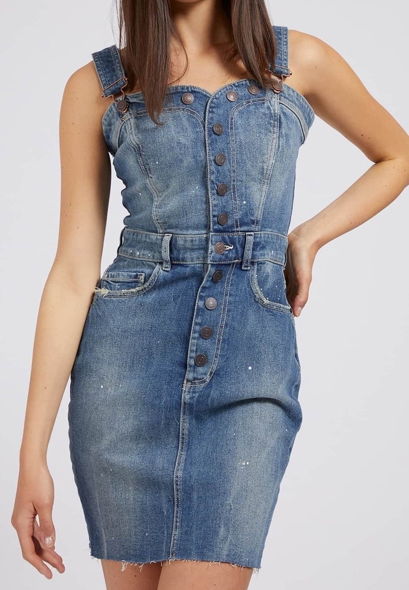 Femme portant une robe en denim bleu sans manches avec des boutons sur le devant, des passants de ceinture et des poches avant, debout avec une main sur la hanche.