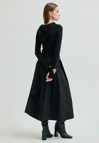 Kobieta o jasnobrązowych włosach, ubrana w czarną bluzę z kapturem, długą czarną spódnicę i czarne botki na obcasach, stoi z rękami złożonymi za plecami.