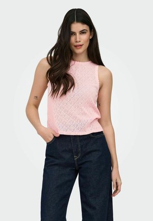 Kobieta z długimi ciemnymi włosami, ubrana w różowy sweterek na ramiączkach i ciemnoniebieskie dżinsy, stoi trzymając jedną rękę w kieszeni.