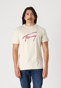 T-shirt en coton beige à manches courtes, avec un logo "Tommy Jeans" rose et bleu imprimé sur la poitrine. Un pantalon en denim visible en dessous.