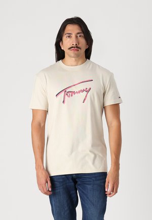 Beige cotton t-shirt with short sleeves, featuring a pink and blue "Tommy Jeans" logo printed across the chest. Denim jeans visible below.