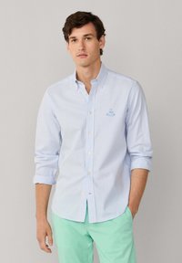 Chemise boutonnée rayée bleu clair en coton, avec un petit logo de voilier brodé sur la poitrine et des manches retroussées.