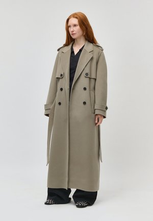 Femme aux cheveux roux portant un long trench-coat beige sur une tenue noire, debout devant un fond gris clair uni.