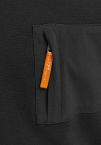 Svart ficka på plagget med en orange etikett. Texturen är slät med ett vävt mönster synligt på det omgivande tyget.