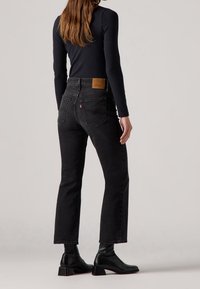 Mujer vestida con jeans rectos de tiro alto negros y botines de tacón negros, de pie contra un fondo claro y liso.
