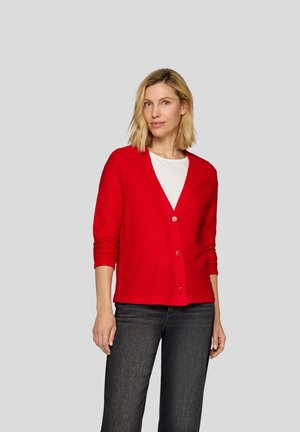 Roter gerippter Cardigan mit V-Ausschnitt, langen Ärmeln und vorderem Knopfverschluss. Getragen über einem weißen Hemd und kombiniert mit dunklen Jeans.