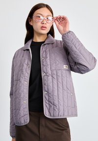 Ανοιχτό μωβ μπουφάν με καπιτονέ ύφασμα, γιακά, κουμπιά με πατ δαχτυλίου και πλαϊνή τσέπη. Φορεμένο πάνω από μαύρο πουκάμισο, στιλιζαρισμένο με ροζ γυαλιά σε σχήμα γάτας.