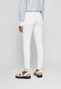 Bílé slim-fit džíny s nenápadnými zadními kapsami, doplněné bílými sandály na pásky. Tkanina se zdá být hladká s jemným leskem.