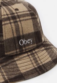 Brown and beige plaid bucket hat made of soft fabric, featuring a white "Obey" logo on the side and a rounded, flexible brim.