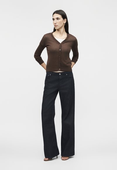 Η γυναίκα στέκεται με τα χέρια πίσω από την πλάτη φορώντας καφέ πλεκτή ζακέτα με κουμπιά πάνω από λευκό τοπ, σκούρο φαρδύ παντελόνι και σανδάλια με τακούνι σε μονόχρωμο φόντο.