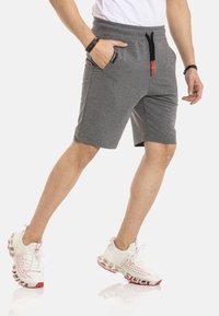 Graue Sportshorts mit einem elastischen Bund, oranges Zugband, Seitentaschen und einer lockeren Passform, kombiniert mit weißen und roten Turnschuhen.