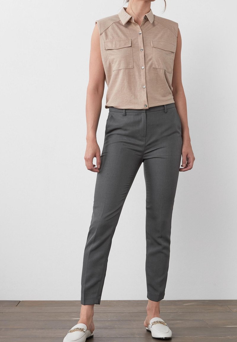 Femme portant une chemise beige sans manches boutonnée, un pantalon gris ajusté et des mocassins blancs à enfiler avec un détail de chaîne dorée.