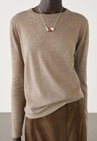 Personne portant un pull beige à manches longues, une jupe en daim marron, et un collier en argent avec des perles multicolores.