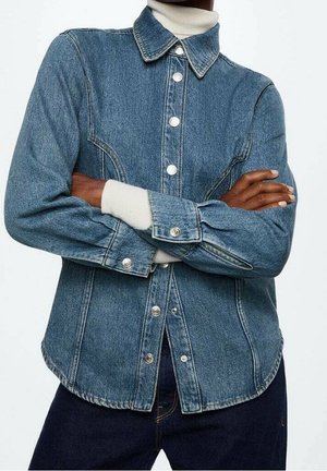 Personne portant une veste en jean bleue sur un col roulé blanc, bras croisés, avec des boutons argentés et un jean bleu foncé visibles.
