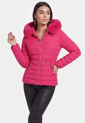 Donna che indossa una giacca imbottita rosa acceso con cappuccio in pelliccia, zip frontale e coulisse in vita, abbinata a pantaloni neri, posa con le mani sui fianchi.