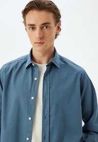 Camisa de botones azul claro con cuello extendido, de tela de algodón y un corte relajado, superpuesta sobre una camiseta blanca de cuello redondo.