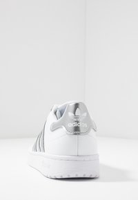 Vit lädersneaker med silverdetaljer, som har en design med tre ränder och en texturerad sula. Bakre delen visar logotypen på en vadderad hälflik.