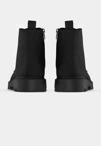 Botas negras hasta el tobillo con un upper sintético liso, cierre de cremallera en la parte posterior y suelas de goma negra gruesa. Diseño de punta redondeada.