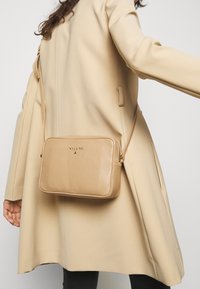 Borsa a tracolla in pelle beige con forma rettangolare, chiusura con zip e un logo discreto. Caratterizzata da una lunga tracolla regolabile e da una texture liscia.