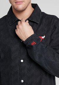 Man som knäpper kragen på svart knäppt jacka med röd Chicago Bulls-logotyp på bröstet och röd emblem på ärmslutet.