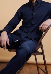 Uomo seduto su una sedia di legno, vestito con una camicia blu scuro e jeans blu, su uno sfondo beige.
