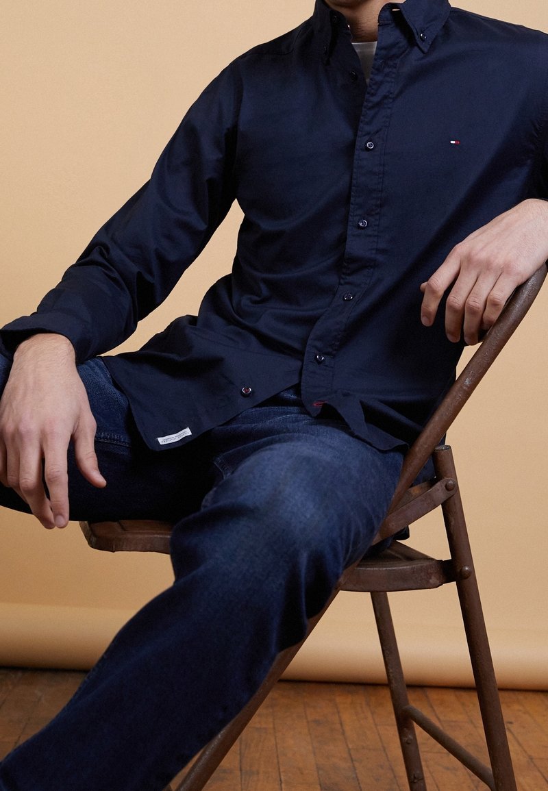 Uomo seduto su una sedia di legno, vestito con una camicia blu scuro e jeans blu, su uno sfondo beige.