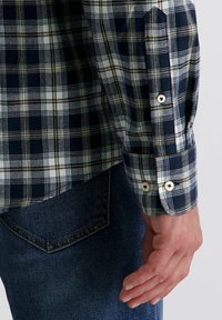 Donkerblauwe geruite overhemd met witte en gele accenten. Heeft knoopmanchetten en een relaxed fit, gecombineerd met donkerdenim jeans.