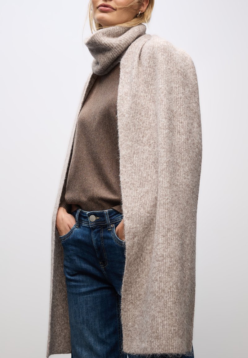Lys beige sjal med en blød, tekstureret strik, båret over en brun sweater. Kombineret med blå denimjeans. Enkel design, ingen synlig hardware eller mønstre.