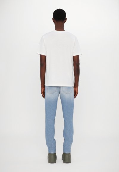 Wei ßes Baumwoll-T-Shirt mit kurzen Ärmeln, kombiniert mit hellblauen Jeans, die am unteren Ende zu einem dunkleren Farbton auslaufen, und grünen Gummistiefeln.