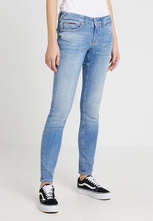 Personne portant un jean slim bleu clair et des baskets montantes noires et blanches, debout devant un fond clair uni.