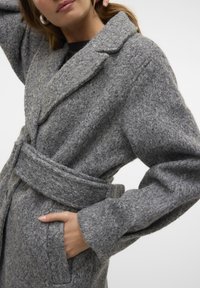Manteau en mélange de laine gris avec une finition texturée, col châle, ceinture à la taille et poches latérales. Présente une silhouette décontractée et une fermeture à boutons.
