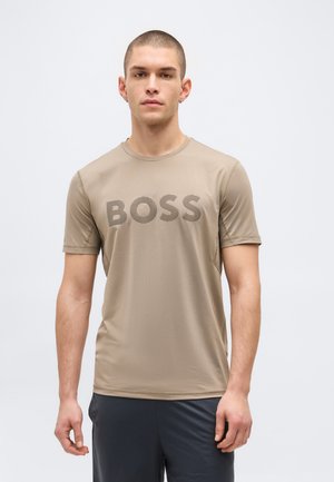 Mladý muž s krátkými vlasy, oblečený v béžovém tričku s nápisem "BOSS" tmavou barvou, v kombinaci s tmavými kalhotami, stojící před jednobarevným pozadím.