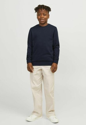 Un pull ras du cou bleu marine, un pantalon beige et des baskets blanches sur un enfant se tenant contre un fond clair. Le tissu semble lisse et décontracté.