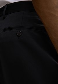 Schwarze Hose mit Knopfverschluss, flacher Vorderseite und einer einzelnen Gesäßtasche in typischer Webstruktur.