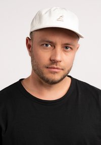 Cappellino bianco in cotone con logo a kangaroo sulla parte frontale, visiera curva e cinturino regolabile. Indossato con una t-shirt nera a girocollo.