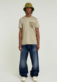 Beige katoenen t-shirt met een camouflagemotief op de borstzak, gecombineerd met loszittende donkerm blauwe denim jeans en witte sneakers. Hoed in emmerstijl inbegrepen.