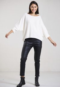 Weißer übergroßer Strickpullover mit weitem Ausschnitt, kombiniert mit schwarzen eng anliegenden Kunstlederhosen und schwarzen knöchelhohen Stiefeln.