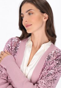 Růžový kardigan s rukávy zdobenými flitry, ribkovou texturou a hlubokým výstřihem do V, vrstvený přes krémovou halenku. Hladká tkanina a měkký střih.