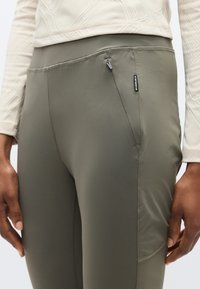 Ελιά πράσινο αθλητικό κολάν με λεία υφή, διαθέτει ασφαλή τσέπη με φερμουάρ και διακριτικές ραφές για εφαρμοστή εμφάνιση.