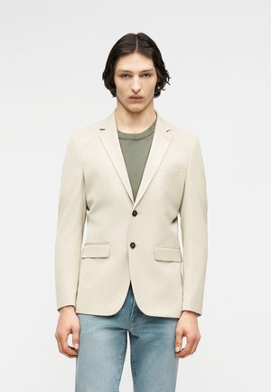 Jeune homme aux cheveux mi-longs noirs portant un blazer beige sur une chemise vert olive et un jean bleu clair, debout devant un fond uni.