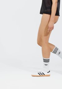 White Adidas shoes with black stripes, paired with knee-high white socks featuring black stripes and logo. Black sheer top, visible legs.