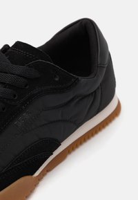 Fekete sneaker sima nejlon és textúrált velúr keverékből, gumitalppal, fűzőzárral és diszkrét logó részletekkel.