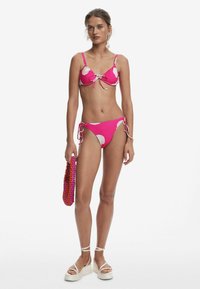 Roze bikini met wit bloemenpatroon, voorzien van verstelbare strikken, gecombineerd met een kleurrijke kralentas en witte platform sandalen.