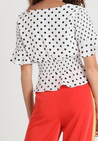 Mujer vistiendo una blusa blanca con lunares y mangas con volantes, y pantalones rojos con franjas amarillas a los lados, vista desde atrás.