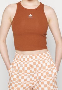 Brązowy prążkowany krótki top z okrągłym dekoltem i białym logo Adidas, zestawiony z pomarańczowo-białymi spodniami w kratkę z wzorem.