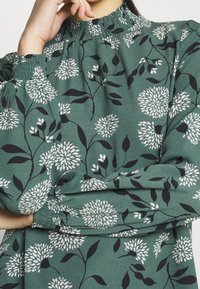 Green floral blouse featuring white flower patterns and black leaves, with gathered cuffs and a ruffled neckline for a textured finish.