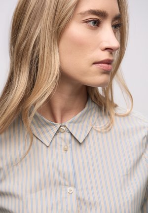 Beżowa koszula na guziki z niebieskimi pionowymi paskami, z klasycznym kołnierzykiem i gładką fakturą, otoczona luźnymi, falującymi włosami.